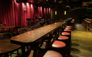 A dimly lit bar with red curtains, long wooden tables, and chairs with red seats. Musical instruments are visible in the background.